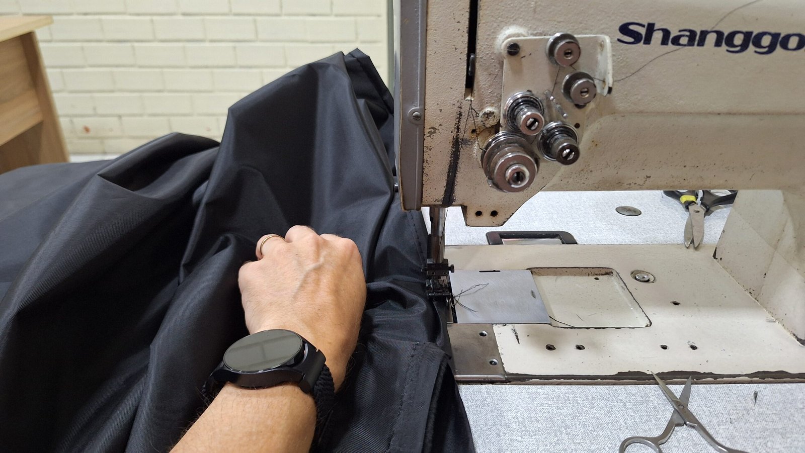 Detalhe de uma máquina de costura industrial trabalhando no acabamento de um tecido preto resistente, representando a fabricação própria das Capas Shanna