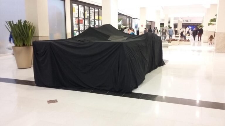 Capa protetora preta em lona instalada em um quiosque de grande porte num corredor de shopping com alta circulação. Padrão estético discreto e profissional para a operação comercial.